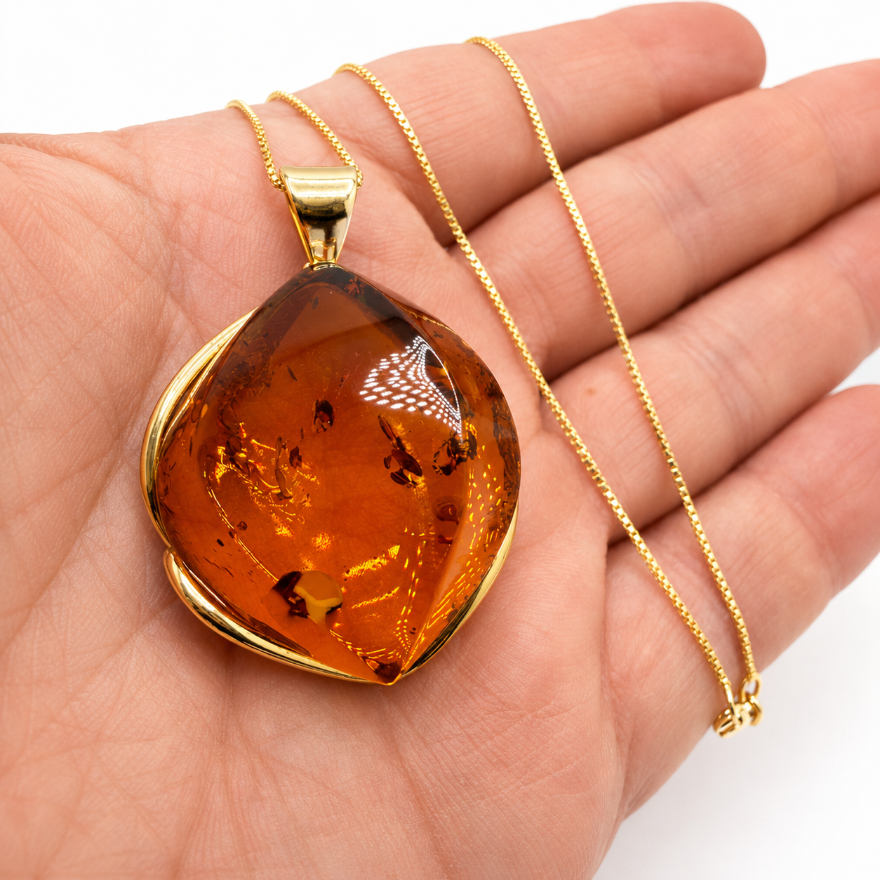 A gold pendant necklace with a large, teardrop-shaped amber gemstone held in a person's hand. The pendant features a polished gold border and a matching gold chain.