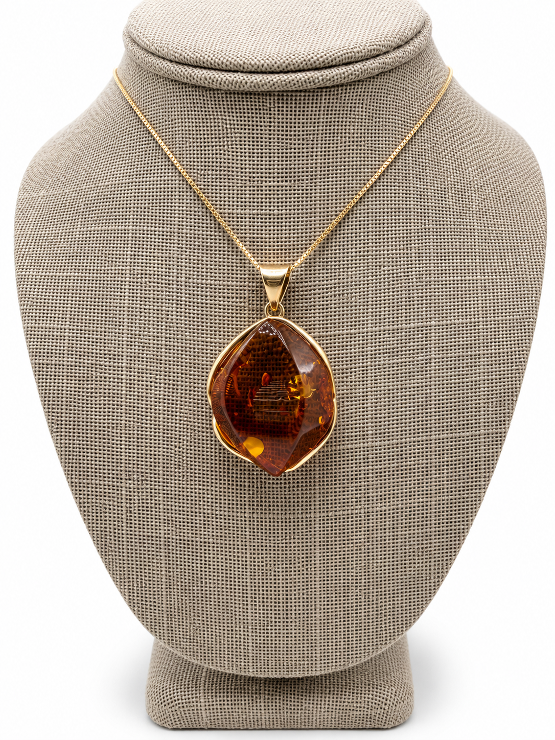A gold pendant necklace featuring a large, organic-shaped amber gemstone, displayed on a textured beige fabric jewelry bust against a white background.